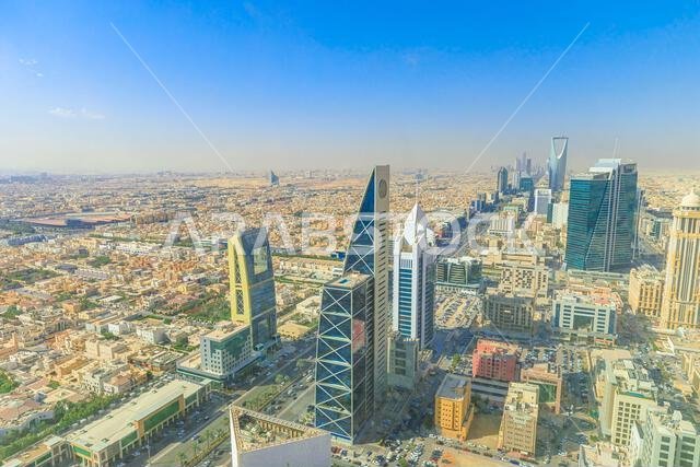 منظر بانورامي لمدينة الرياض، المملكة العربية السعودية، يُظهر امتدادها الحضري الشاسع، وشوارعها الصاخبة، وهندستها المعمارية الحديثة تحت سماء مشرقة مشمسة.