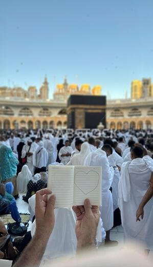 حاج يحمل دفتر ملاحظات صغيرًا عليه رسمة قلب أمام الكعبة المشرفة في مكة المكرمة بالمملكة العربية السعودية. الموقع المقدس محاط بالمصلين