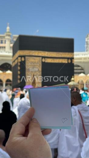 حاج يحمل دفتر ملاحظات صغيرًا عليه رسمة قلب أمام الكعبة المشرفة في مكة المكرمة بالمملكة العربية السعودية. الموقع المقدس محاط بالمصلين