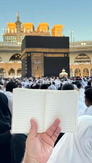 حاج يحمل دفتر ملاحظات صغيرًا عليه رسمة قلب أمام الكعبة المشرفة في مكة المكرمة بالمملكة العربية السعودية. الموقع المقدس محاط بالمصلين