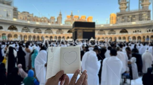 حاج يحمل دفتر ملاحظات صغيرًا عليه رسمة قلب أمام الكعبة المشرفة في مكة المكرمة بالمملكة العربية السعودية. الموقع المقدس محاط بالمصلين