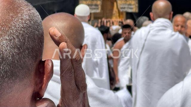 A group of Muslim pilgrims dressed in white ihram garments performing Hajj near the Kaaba in Mecca, Saudi Arabia. A close-up of a man raising his hand
