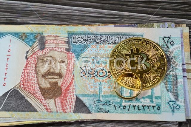 Saudi Arabian money banknotes bills of SAR riyals and Bitcoin physical golden coin, BTC the 1st decentralized cryptocurrency and a gold ring pure yell