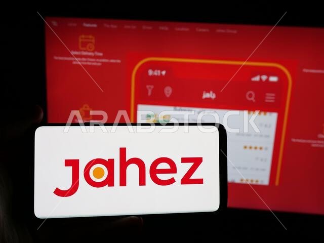 شتوتغارت، ألمانيا - 13-06-2025: شخص يحمل هاتفًا ذكيًا عليه شعار شركة جاهز السعودية لتطبيقات توصيل الطعام على الشاشة أمام موقع الويب.
