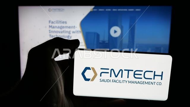 Stuttgart, Germany - 06-14-2025: Person holding mobile phone with logo of Saudi Facility Management Company (FMTECH) on screen in front of web page.