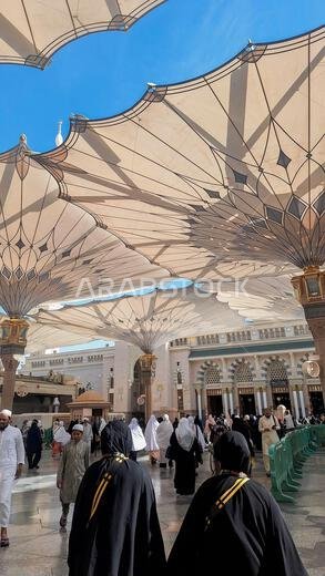 Medina, Saudi Arabia - March 6, 2024:  Pilgrims walk to the main mosque of the Prophet Muhammad Al-Masjid an-Nabawi underneath giant umbrella preparin