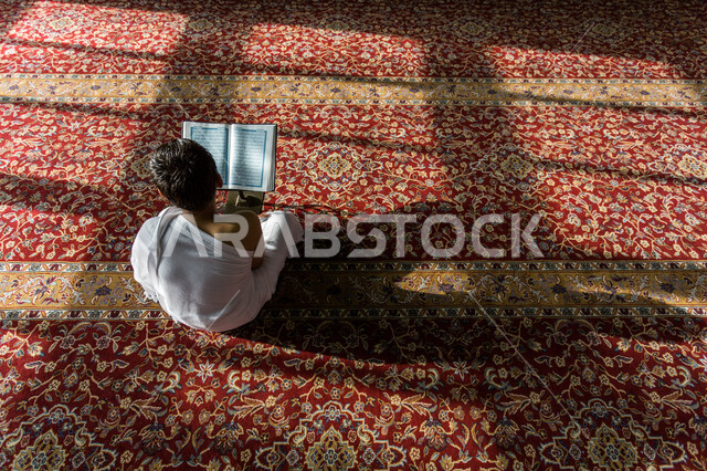 صورة من الخلف لولد عربي خليجي يرتدي ملابس الاحرام ويجلس في المسجد وامامه المصحف الشريف على حامل خشبي، الصلاه والدعاء في المسجد، العبادة والتقرب الى الله، قراءة وتلاوة القران، التعبد في بيوت الله، ذكر الله، تحفيظ القران الكريم، تفسير القرآن