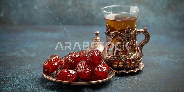 إفطار رمضاني كريم مع التمر المجفف، وجبة إفطار تتضمن التمر والشاي العربي في كوب تقليدي، منظر من زاوية مختلفة على خلفية زرقاء ريفية.