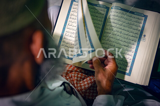 A picture from the back of an old man holding the Holy Qur’an in his hand, praying and supplicating in the mosque, worshiping and getting closer to God, reading and reciting the Qur’an, worshiping in the homes of God, remembrance of God, memorizing the Ho