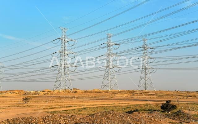 أبراج نقل كهرباء فولاذية ضخمة مزودة بكابلات، وخلفية من السماء الزرقاء، وشمس الظهيرة تسطع على منظر طبيعي صحراوي إلى حد ما.