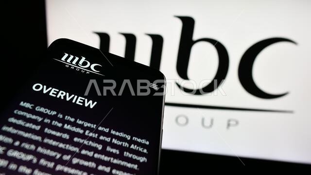 هاتف محمول يعرض موقع مجموعة إم بي سي الإعلامية السعودية أمام شعار الشركة. التركيز على أعلى يسار شاشة الهاتف. هاتف محمول يعرض موقع مجموعة إم بي سي الإعلامية السعودية أمام شعار الشركة. التركيز على أعلى يسار شاشة الهاتف.