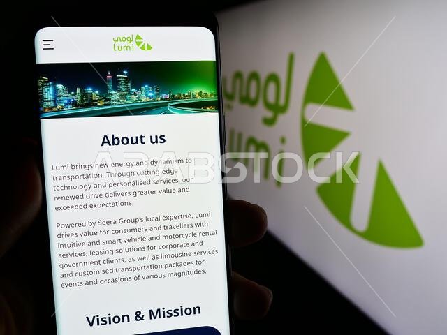 شخص يحمل هاتفًا ذكيًا يعرض صفحة ويب لشركة تأجير السيارات السعودية "لومي" أمام الشعار. التركيز على منتصف شاشة الهاتف. شخص يحمل هاتفًا ذكيًا يعرض صفحة ويب لشركة تأجير السيارات السعودية "لومي" أمام الشعار. التركيز على منتصف شاشة الهاتف.