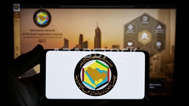 شخص يحمل هاتفًا ذكيًا عليه شعار الاتحاد العربي ومجلس التعاون الخليجي أمام موقع إلكتروني. التركيز على شاشة الهاتف.