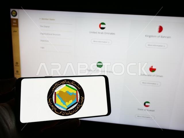 شخص يحمل هاتفًا محمولًا عليه شعار الاتحاد العربي ومجلس التعاون الخليجي (GCC) أمام صفحة ويب. التركيز على شاشة الهاتف.