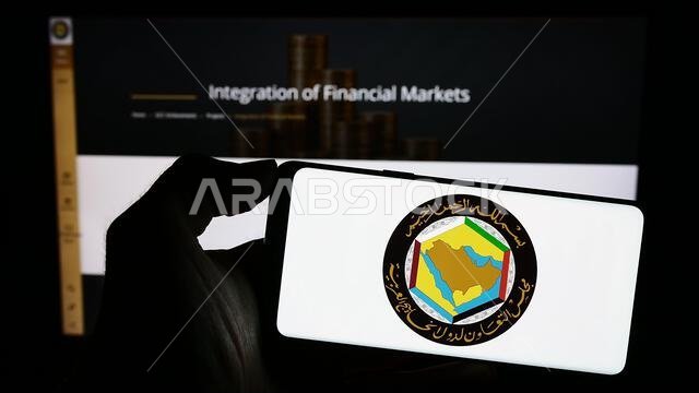 شخص يحمل هاتفًا محمولًا عليه شعار الاتحاد العربي ومجلس التعاون الخليجي أمام صفحة ويب. التركيز على شاشة الهاتف.