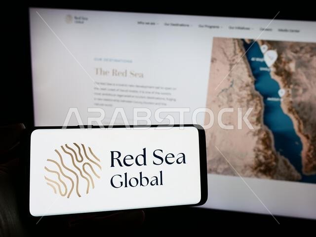 شخص يحمل هاتفًا محمولًا عليه شعار شركة التطوير العقاري السعودية "البحر الأحمر العالمي" أمام صفحة ويب خاصة بالشركة. التركيز على شاشة الهاتف. شخص يحمل هاتفًا محمولًا عليه شعار شركة التطوير العقاري السعودية "البحر الأحمر العالمي" أمام صفحة ويب خاصة بالشركة. التركيز على شاشة الهاتف.