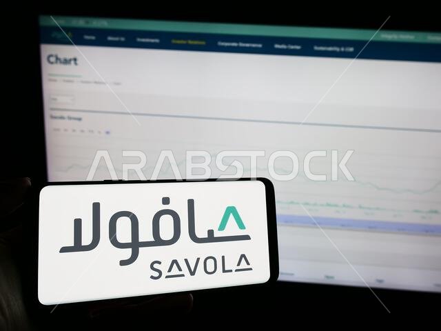 شخص يحمل هاتفًا محمولًا عليه شعار شركة سافولا جروب الصناعية السعودية، أمام صفحة ويب خاصة بالشركة. التركيز على شاشة الهاتف.