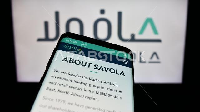 Mobile phone with website of Saudi Arabian industrial company Savola Group in front of business logo. Focus on top-left of phone display.