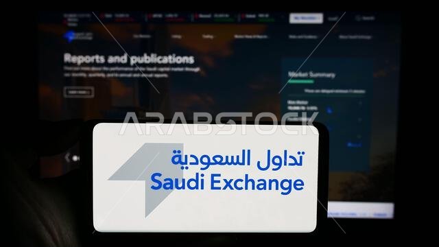 شخص يحمل هاتفًا محمولًا عليه شعار البورصة العربية السعودية (تداول) أمام صفحة ويب خاصة بالشركة. التركيز على شاشة الهاتف. شخص يحمل هاتفًا محمولًا عليه شعار البورصة العربية السعودية (تداول) أمام صفحة ويب خاصة بالشركة. التركيز على شاشة الهاتف.