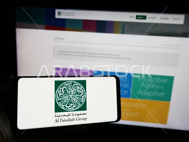 شخص يحمل هاتفًا محمولًا عليه شعار شركة الفيصلية السعودية أمام صفحة ويب خاصة بالشركة. التركيز على شاشة الهاتف. شخص يحمل هاتفًا محمولًا عليه شعار شركة الفيصلية السعودية أمام صفحة ويب خاصة بالشركة. التركيز على شاشة الهاتف.