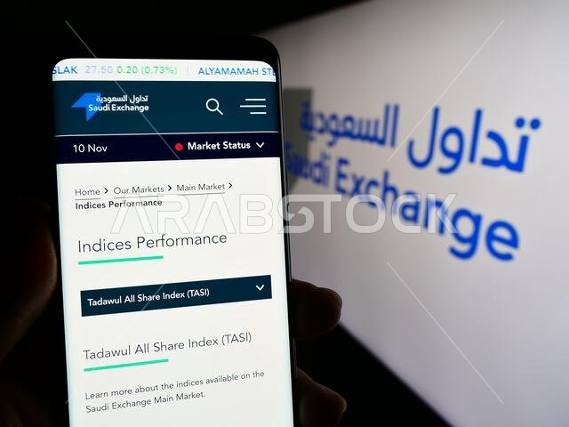 شخص يحمل هاتفًا ذكيًا يعرض صفحة ويب لبورصة السعودية (تداول) أمام الشعار. التركيز على منتصف شاشة الهاتف. شخص يحمل هاتفًا ذكيًا يعرض صفحة ويب لبورصة السعودية (تداول) أمام الشعار. التركيز على منتصف شاشة الهاتف.