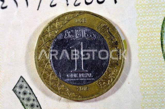 الوجه الخلفي لعملة معدنية من فئة ريال سعودي واحد لعام 2016 على خلفية ورقة نقدية من فئة ريال سعودي واحد، ريال معدني واحد وورقة نقدية من فئة ريال سعودي معًا، السعودية