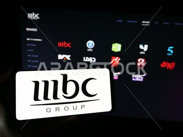 شخص يحمل هاتفًا ذكيًا عليه شعار مجموعة إم بي سي الإعلامية السعودية، يقف أمام موقع إلكتروني. التركيز على شاشة الهاتف.