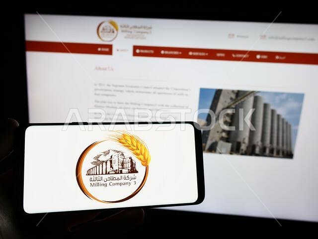 شخص يحمل هاتفًا ذكيًا عليه شعار شركة الطحن السعودية رقم 3 (MC3) على الشاشة أمام موقع إلكتروني. التركيز على شاشة الهاتف. شخص يحمل هاتفًا ذكيًا عليه شعار شركة الطحن السعودية رقم 3 (MC3) على الشاشة أمام موقع إلكتروني. التركيز على شاشة الهاتف.