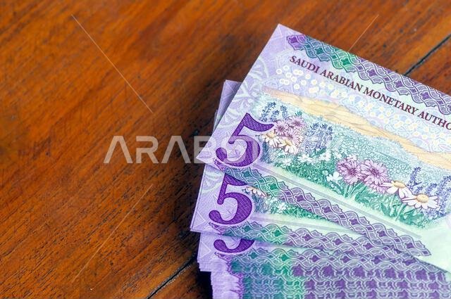 كومة من أوراق نقدية من فئة خمسة ريالات سعودية موضوعة على طاولة خشبية، مع التركيز على الصورة المختارة