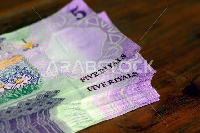 كومة من أوراق نقدية من فئة خمسة ريالات سعودية موضوعة على طاولة خشبية، مع التركيز على الصورة المختارة