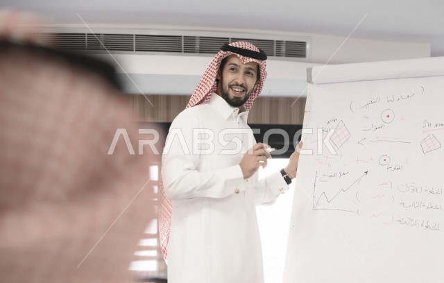 صورة مقربة لمدرب سعودي خليجي مبتسم يشرح على لوح ابيض  ، رجل اعمال في اجتماع لشرح استراتيجيات الشركة ، غرفة اجتماعات ، اجتماع عمل ، دورة تدريبية