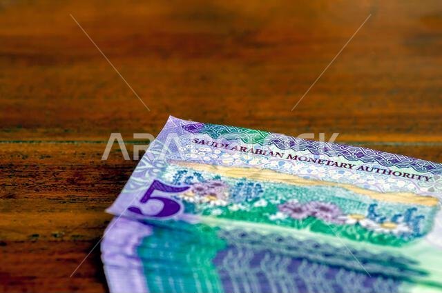 كومة من أوراق نقدية من فئة خمسة ريالات سعودية موضوعة على طاولة خشبية، مع التركيز على الصورة المختارة
