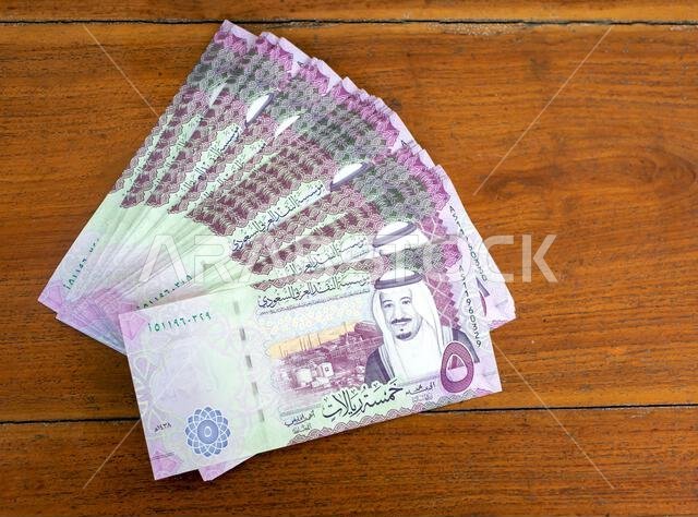 كومة من أوراق نقدية من فئة خمسة ريالات سعودية موضوعة على طاولة خشبية، مع التركيز على الصورة المختارة