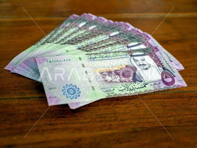 كومة من أوراق نقدية من فئة خمسة ريالات سعودية موضوعة على طاولة خشبية، مع التركيز على الصورة المختارة