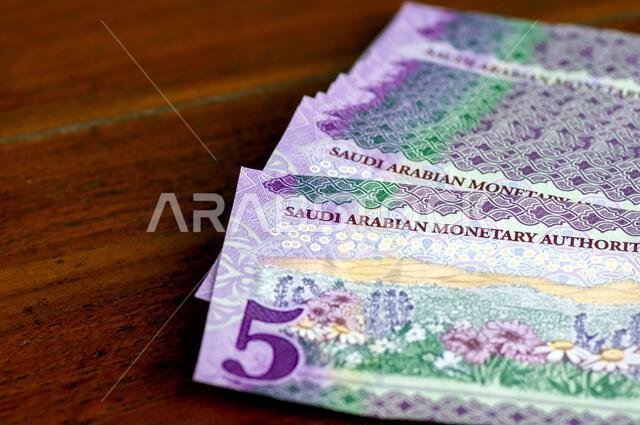 كومة من أوراق نقدية من فئة خمسة ريالات سعودية موضوعة على طاولة خشبية، مع التركيز على الصورة المختارة
