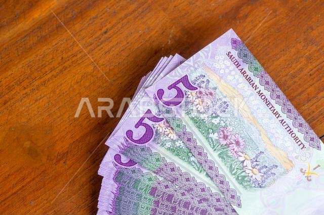كومة من أوراق نقدية من فئة خمسة ريالات سعودية موضوعة على طاولة خشبية، مع التركيز على الصورة المختارة