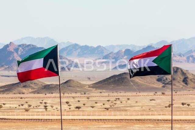 أعلام الكويت والسودان ترفرف معاً في مهب الريح في صحراء المملكة العربية السعودية، مع وجود الجبال في الخلفية، العلا، المملكة العربية السعودية