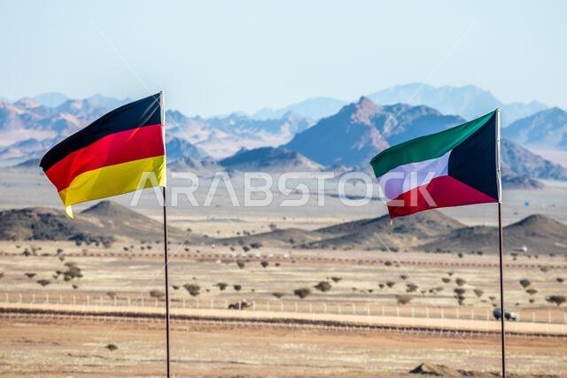 أعلام ألمانية وكويتية ترفرف معاً في مهب الريح في صحراء المملكة العربية السعودية، مع وجود الجبال في الخلفية، العلا، المملكة العربية السعودية