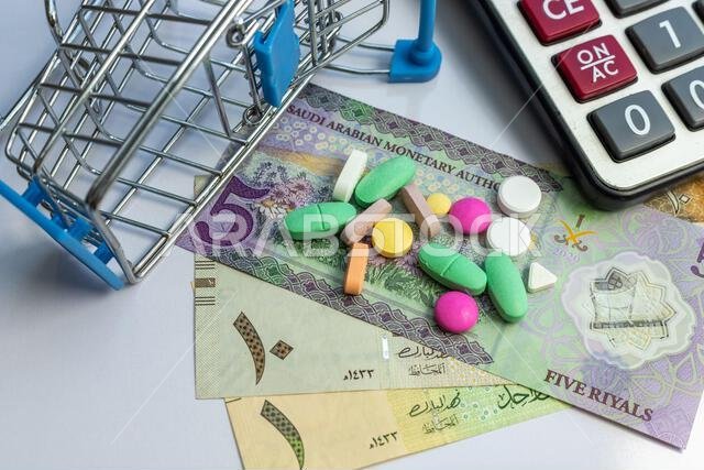 A variety of pills in spilling from a mini shopping trolley on a Saudi Arab banknotes. Health and medicine cost concept in KSA A variety of pills in spilling from a mini shopping trolley on a Saudi Arab banknotes. Health and medicine cost concept in KSA