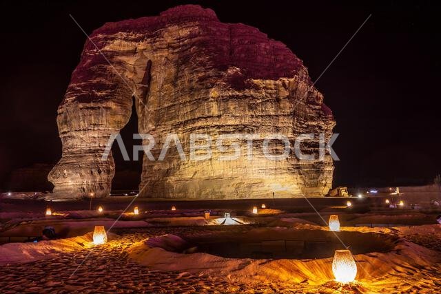 صالة خارجية مضاءة أمام صخرة الفيل المتآكلة، الواقعة في صحراء العلا ليلاً، المملكة العربية السعودية
