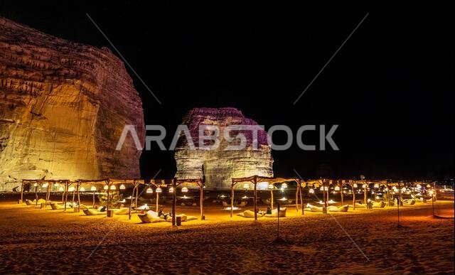 صالة خارجية مضاءة أمام صخرة الفيل المتآكلة، الواقعة في صحراء العلا ليلاً، المملكة العربية السعودية
