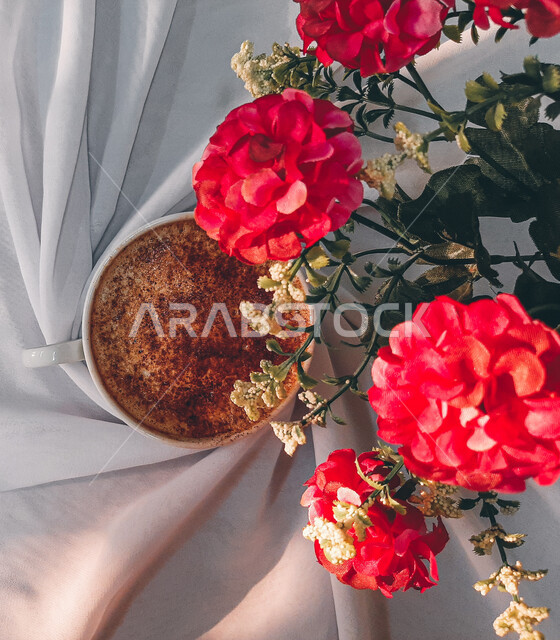 صورة مقربة لفنجان القهوة فوق غطاء ابيض اللون، مشروب الكابتشينو الساخن، قهوة اسبريسو، مشروبات ساخنة لذيذة، فنجان القهوة الساخن برغوة كثيفة، ازهار الزينة الحمراء