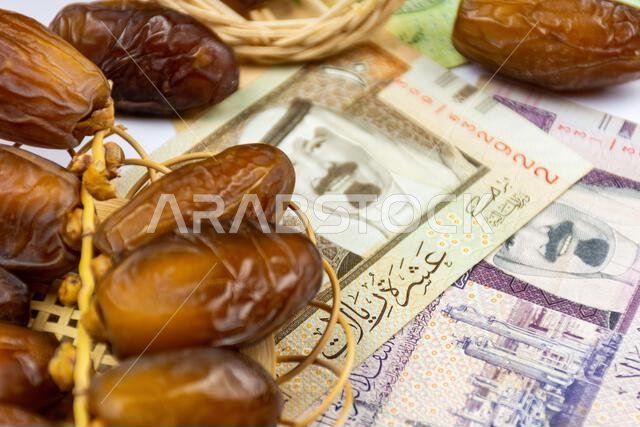 صورة مقرّبة لتمور جزائرية ملكية على طبق خشبي. عملات ورقية سعودية. مفهوم رمضان.