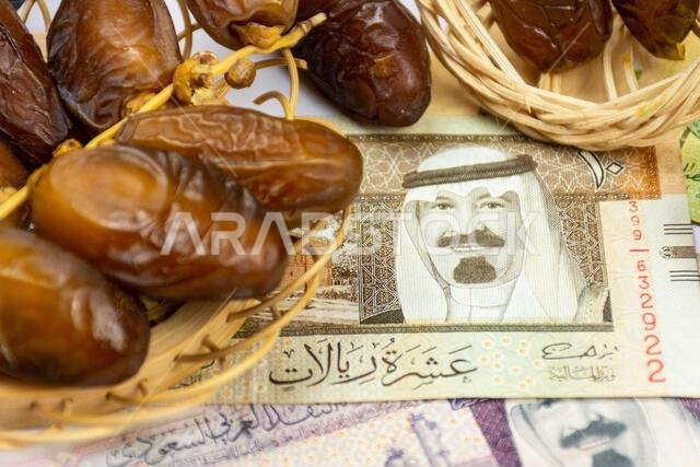 صورة مقرّبة لتمور جزائرية ملكية على طبق خشبي. عملات ورقية سعودية. مفهوم رمضان.