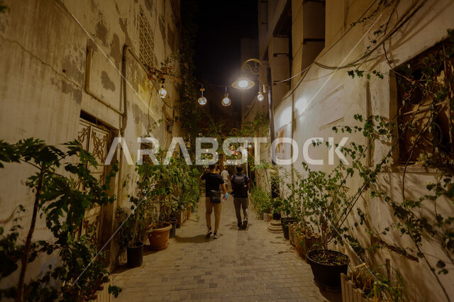 صورة من الخلف لشابين يتجولون في ازقة البلد التاريخي بجدة في المملكة العربية السعودية ليلا، المباني الأثرية، المدينة التاريخية في جدة، التصميم المعماري العربي القديم، أماكن سياحية، المعالم التاريخية في جدة