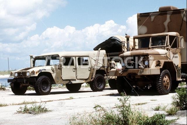 مركبة هامفي متعددة الأغراض عالية الحركة من طراز M998 (HMMWV)، على اليسار، وشاحنة وزنها 5 أطنان، تقفان على الرصيف قبل تحميلهما على متن سفينة إنزال. ستنقل السفينة معدات مشاة البحرية الأمريكية إلى سفينة تابعة للبحرية، والتي بدورها ستنقلها إلى المملكة العربية السعودية لدعم عملية درع الصحراء. العملية/السلسلة: درع الصحراء، الولاية: كارولاينا الشمالية، الدولة: الولايات المتحدة الأمريكية.