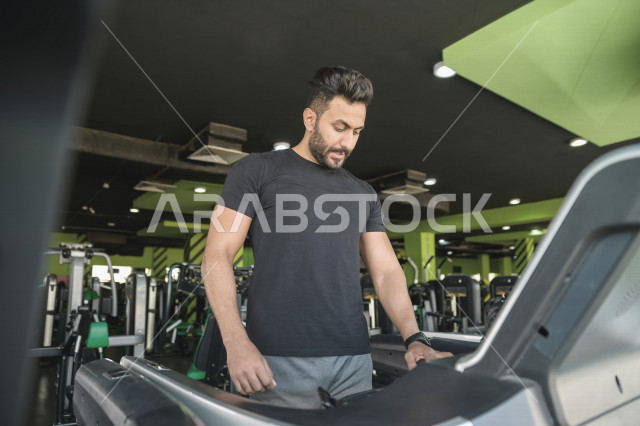 شاب عربي خليجي سعودي يقوم بالتمرن على جهاز المشي ، ممارسة التمارين الرياضية في النادي الرياضي