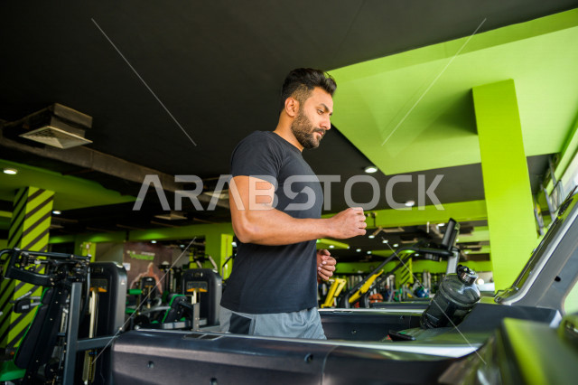 شاب عربي خليجي سعودي يقوم بالتمرن على جهاز المشي ، ممارسة التمارين الرياضية في النادي الرياضي