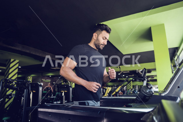 شاب عربي خليجي سعودي يقوم بالتمرن على جهاز المشي ، ممارسة التمارين الرياضية في النادي الرياضي