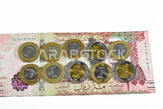 ورقة نقدية من فئة 100 ريال سعودي مع رزمة من العملات المعدنية من فئة ريال واحد وريالين، تحمل صورة الملك سلمان بن عبد العزيز آل سعود.